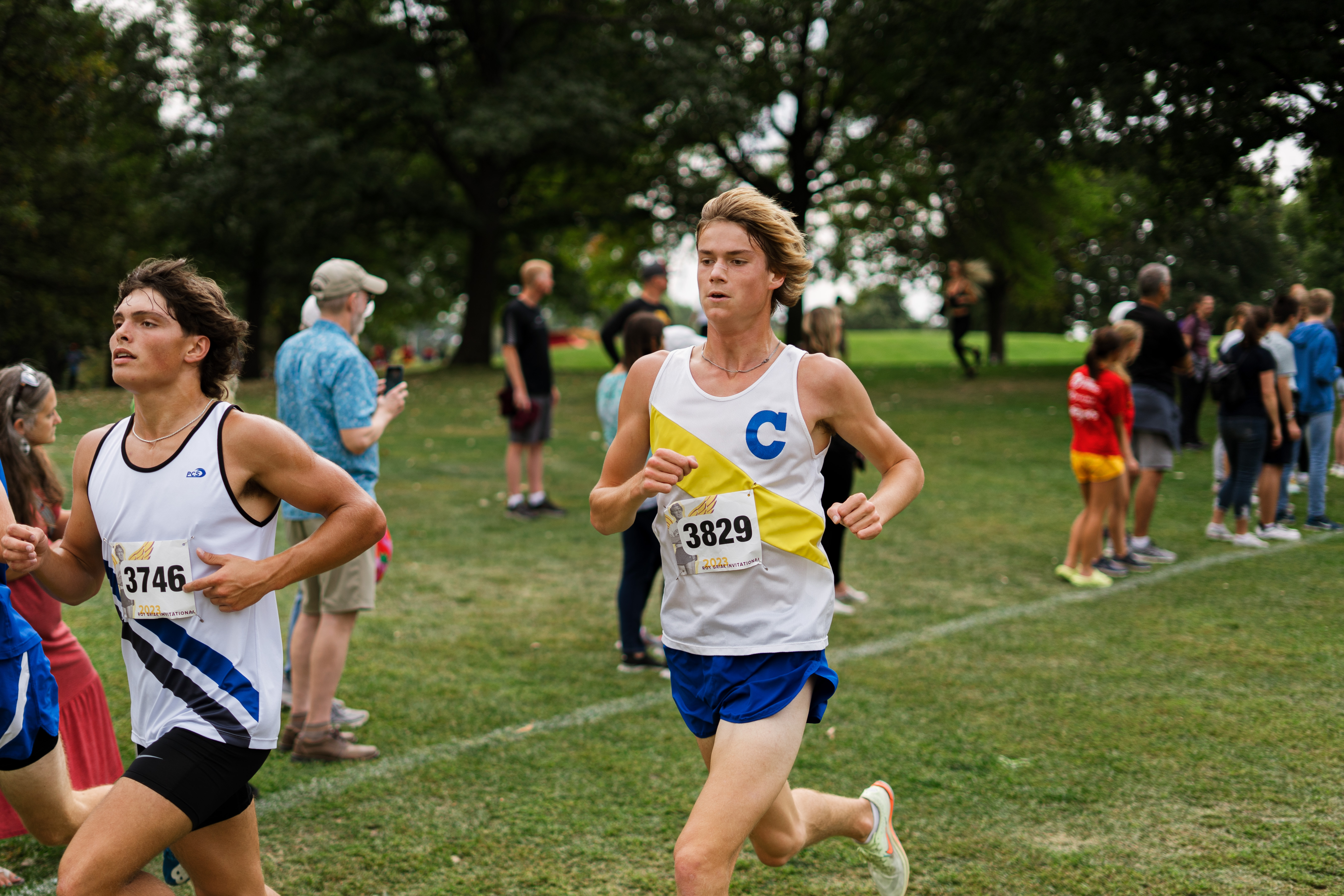 Two young men run a race on a grassy field, other runners and spectators are visible in the background.